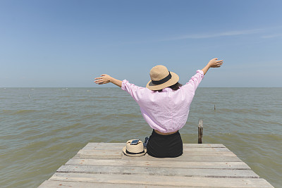 一名女子举起双手，坐在木墩上，面对着大海和蓝天