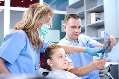 一名专业女牙医在男医生协助下，在牙科诊所检查一名小女孩病人的x光影像。