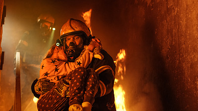 勇敢的消防队员走下燃烧的建筑物的楼梯，将获救的女孩抱在怀里。开火，背景中有一名消防员。