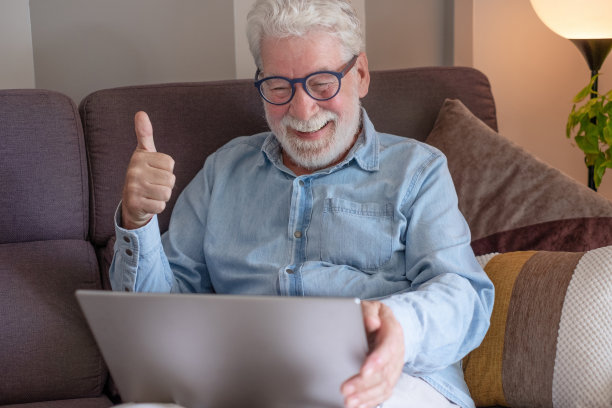 戴眼镜的老人坐在沙发上用笔记本电脑。
